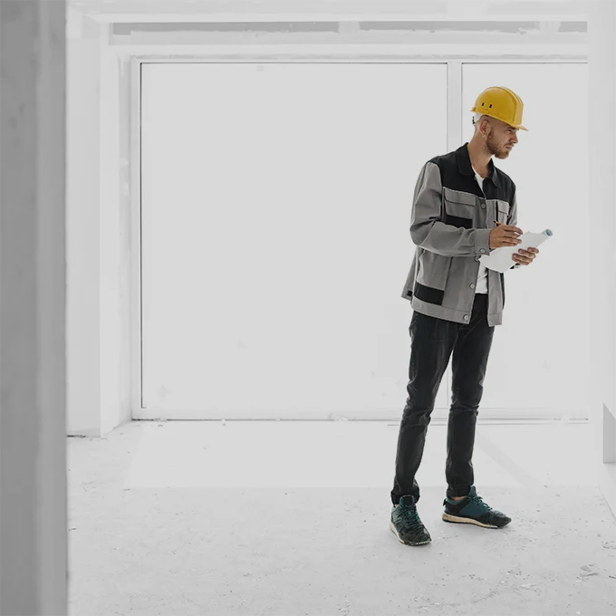 Ein Bauingenieur mit gelbem Helm auf dem Kopf hat Pläne und Stift in der Hand und schaut sich etwas an.